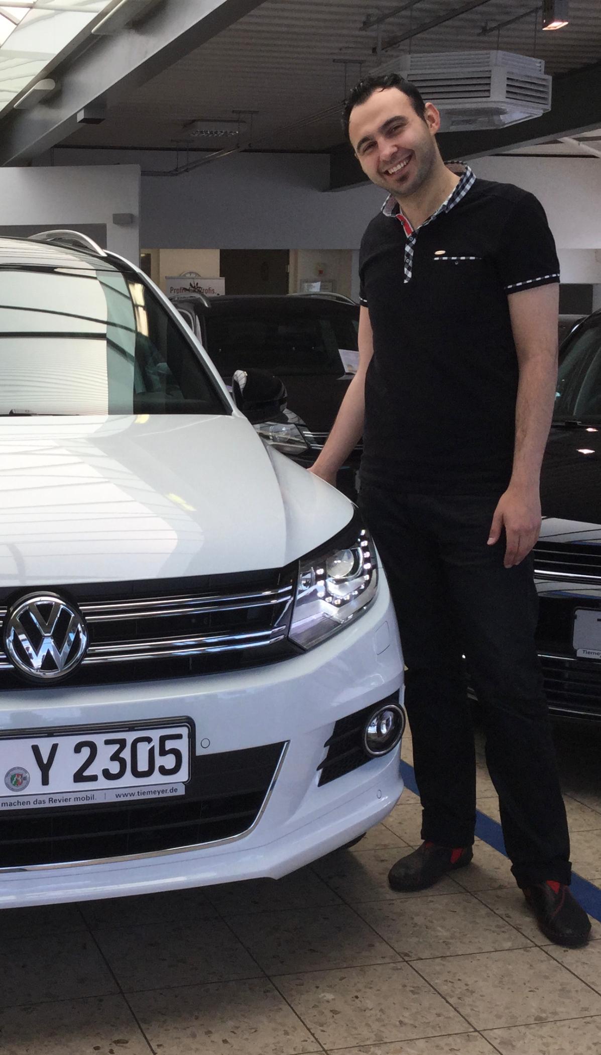 Fahrlehrer Yalcin steht lächelnd neben einem weißen VW Fahrschulauto in Datteln.